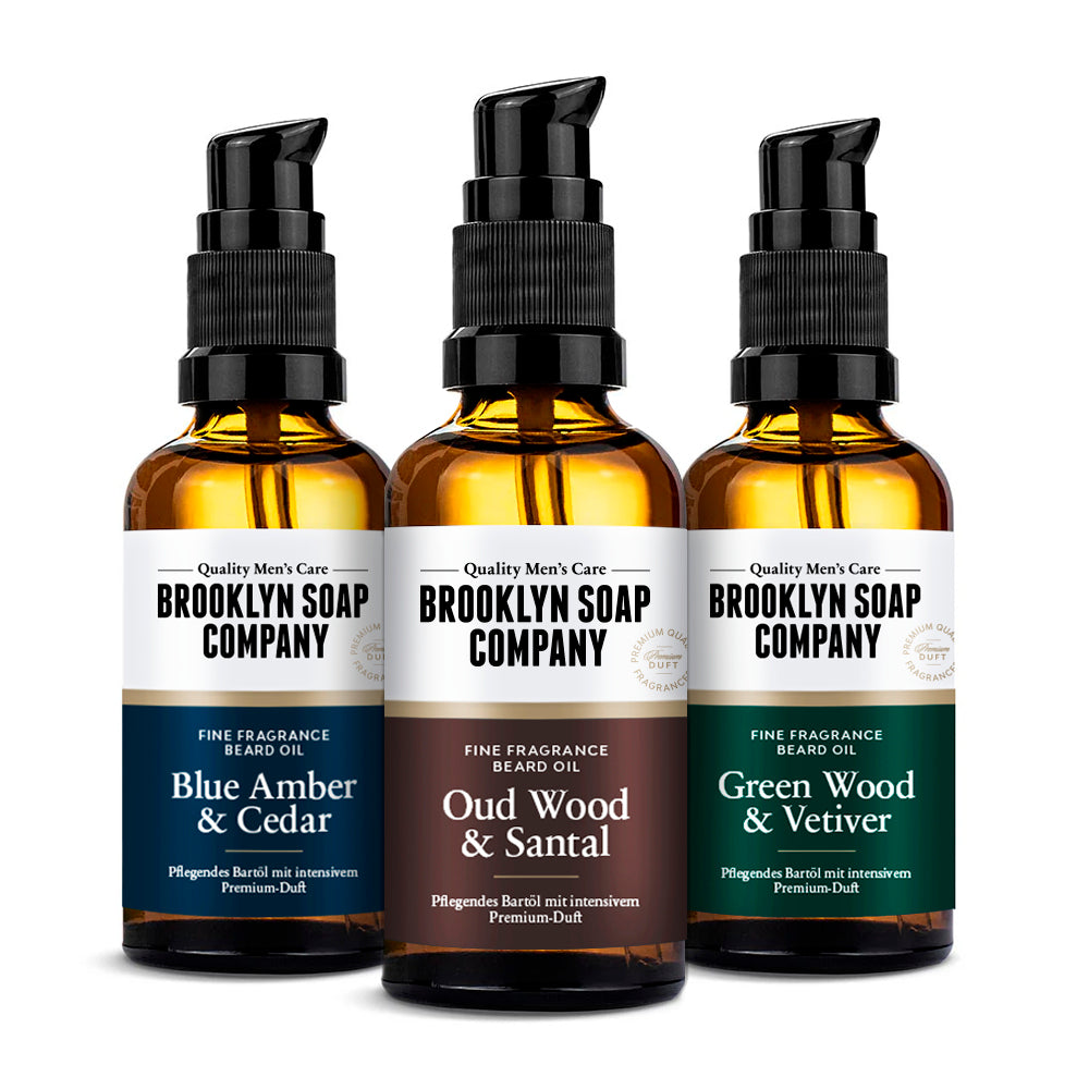 Drei Bartöle der Brooklyn Soap Company – Blue Amber & Cedar, Oud Wood & Santal sowie Green Wood & Vetiver – in bernsteinfarbenen Glasflaschen mit Pumpspender, auf einer Holzoberfläche vor einem sanft verlaufenden hellgrauen Hintergrund