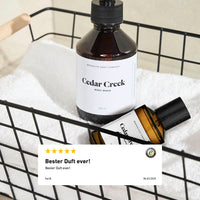 Zwei Produkte aus der Cedar Creek Serie von Brooklyn Soap Company – Body Wash und Eau de Parfum – stehen in einem schwarzen Drahtkorb auf einem weißen Handtuch. Im Vordergrund eine eingeblendete 5-Sterne-Bewertung mit dem Kommentar „Bester Duft ever!“.