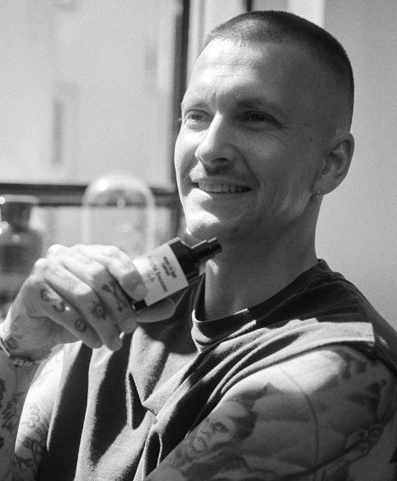 Schwarz-weiß Foto eines lächelnden Mannes mit Tätowierungen, der ein Bartöl-Fläschchen an seine Wange hält.